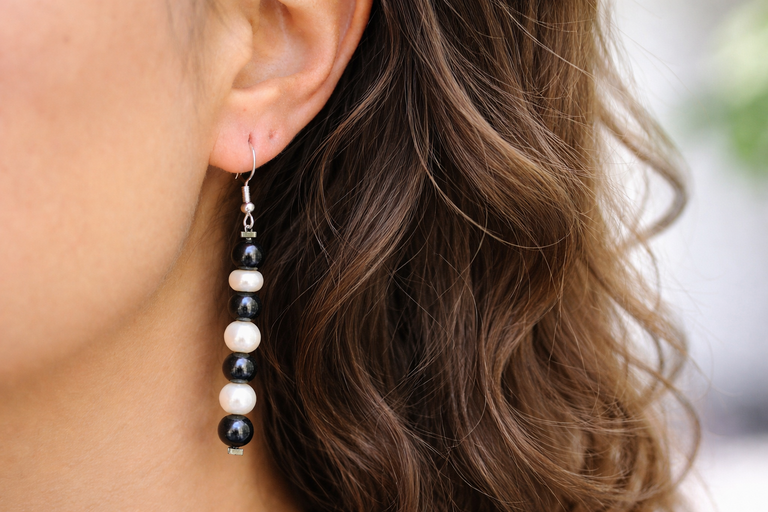Close-up of a woman’s ear wearing long drop earrings made of alternating black shungite beads and white pearls on silver hooks, with softly lit longer wavy brown hair framing the side of her face.