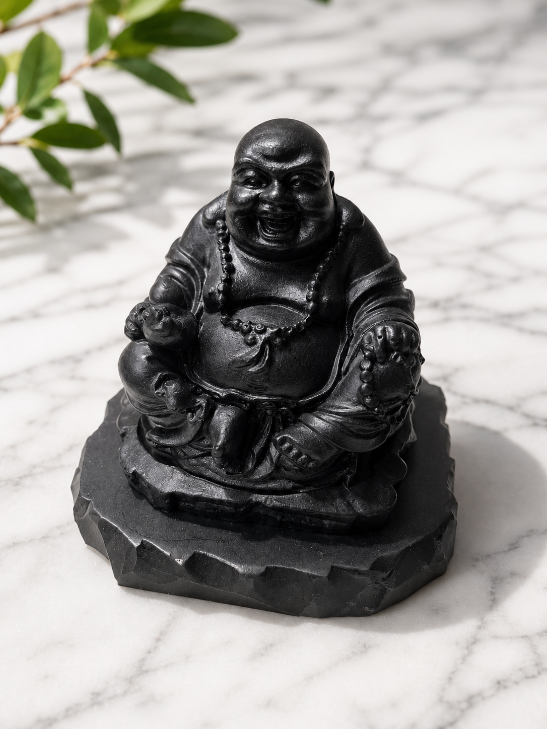 Handcrafted shungite Laughing Buddha statue carved from genuine black shungite stone, displayed on a matching stone base atop a white marble surface. The smiling Buddha features intricate robe details and prayer beads, highlighted by soft natural lighting and gentle shadows, creating a peaceful and grounding decorative accent for meditation spaces, altars, and mindful interiors.