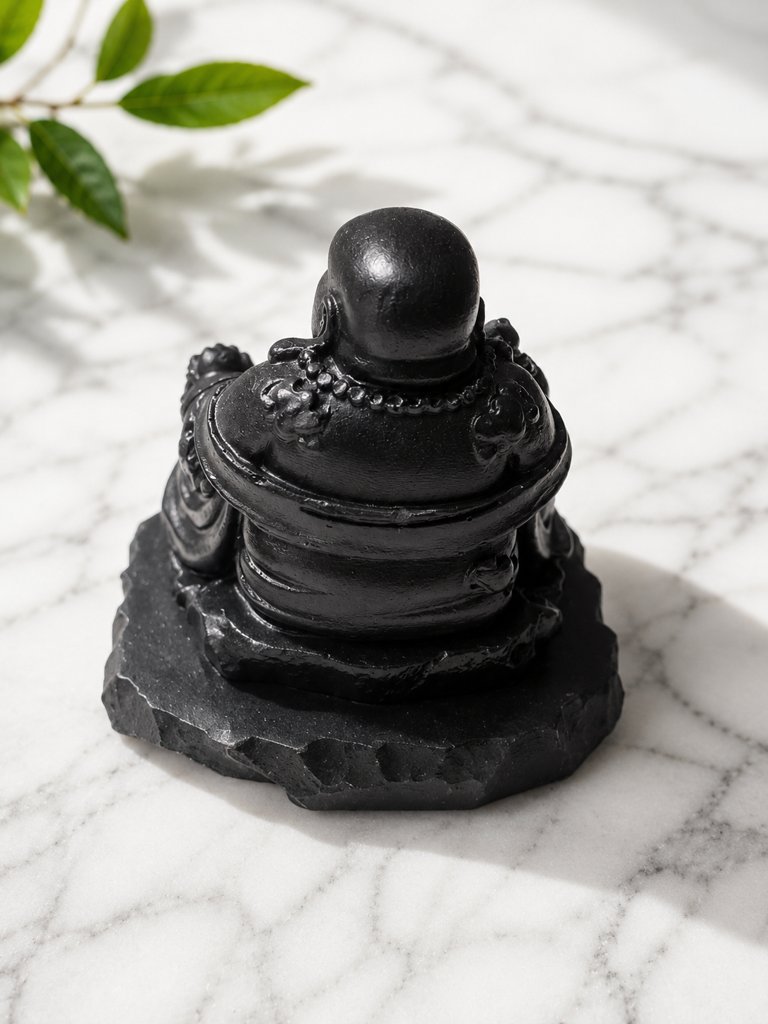 Handcrafted black shungite laughing Buddha statue viewed from the back, seated on a natural shungite stone base. The polished carving features detailed robes and bead necklace accents, displayed on a white marble surface with soft natural lighting and subtle shadows. A decorative spiritual sculpture for meditation spaces, altars, home décor, and mindful living environments.