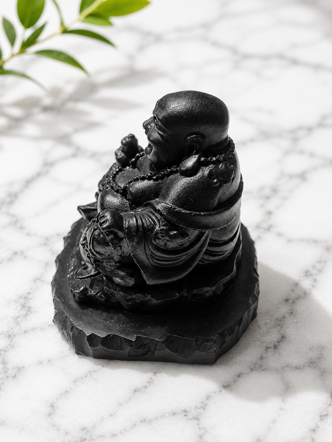Side view of a handcrafted black shungite laughing Buddha statue seated on a natural shungite stone base. The polished carving showcases detailed robes, prayer beads, and smooth contours, displayed on a white marble surface with soft natural lighting and gentle shadows. A decorative spiritual sculpture for meditation spaces, altars, mindful living, and home décor.