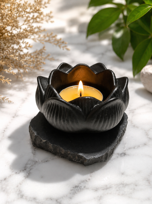 Black shungite lotus candle holder with a lit tea light candle, displayed on a marble surface with soft natural lighting, calming shadows, and botanical accents in a serene spa-inspired setting.