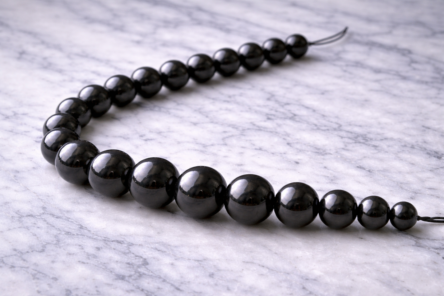 Polished shungite craft beads on string, arranged from small to large on white marble surface, showing glossy black finish in sizes from 6mm to 12mm.