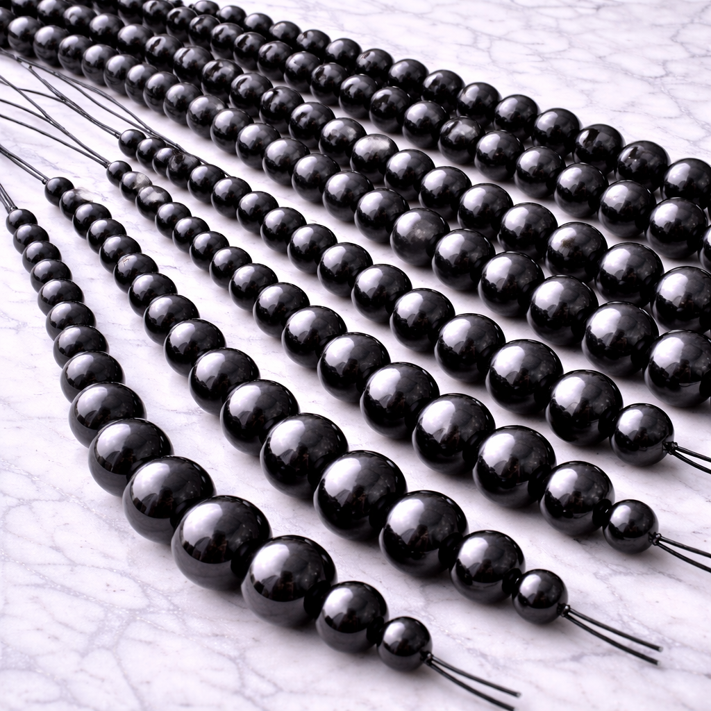 Multiple strands of polished black shungite beads in graduated sizes (approximately 6mm to 12mm) arranged side by side on a white marble surface, showing several bead strings together for craft and jewelry making.