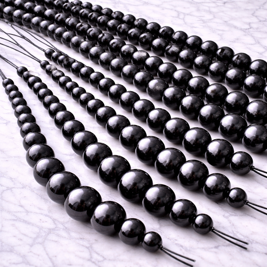 Multiple strands of polished black shungite beads in graduated sizes (approximately 6mm to 12mm) arranged side by side on a white marble surface, showing several bead strings together for craft and jewelry making.