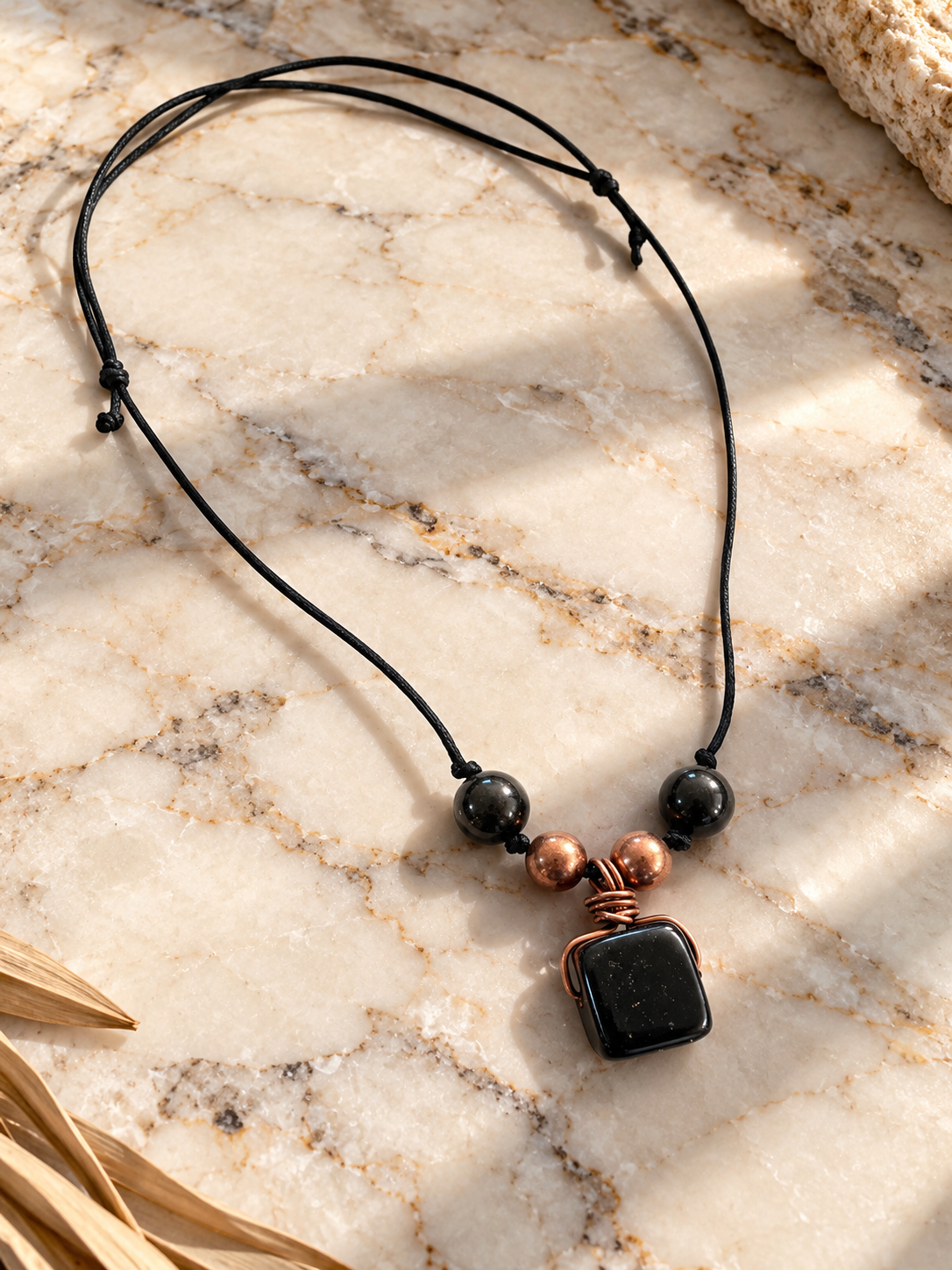 Polished black shungite pendant wrapped in copper wire with copper and shungite beads on an adjustable black cord, displayed on natural cream marble.