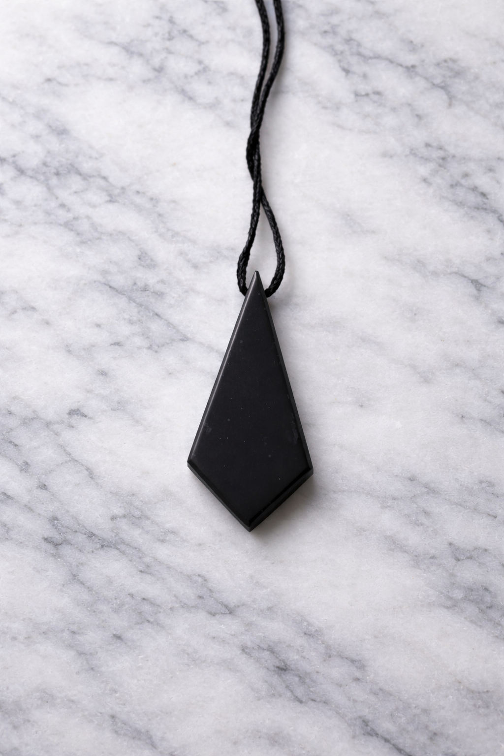 Polished black shungite diamond-shaped pendant on black cord displayed on a white marble surface with soft natural lighting