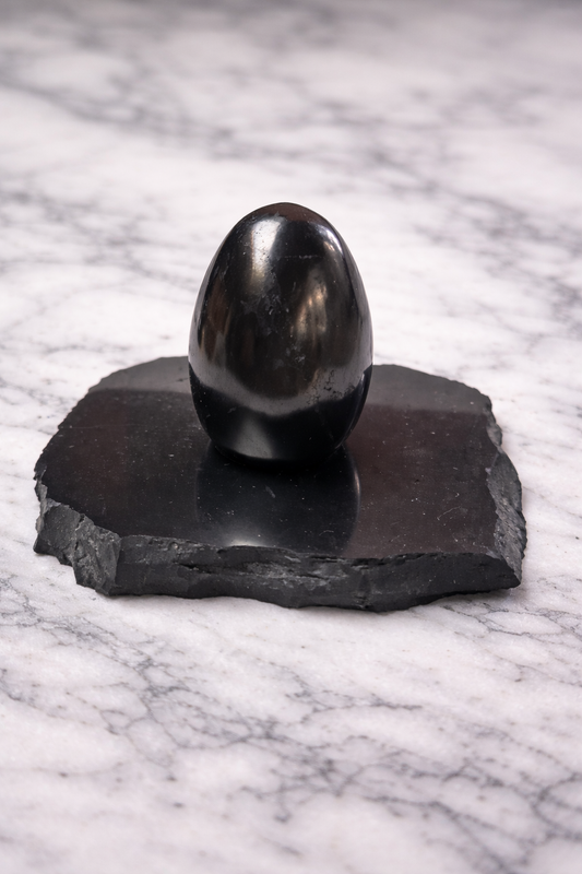 Polished shungite egg crystal on raw shungite base placed on white marble surface with soft natural lighting, highlighting smooth glossy finish and natural stone texture