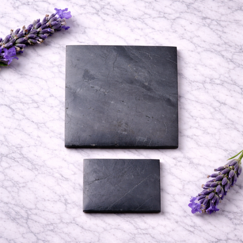 Two polished shungite plates in different square sizes displayed on a white marble surface, accented with purple lavender sprigs, showing natural stone veining and matte black texture.