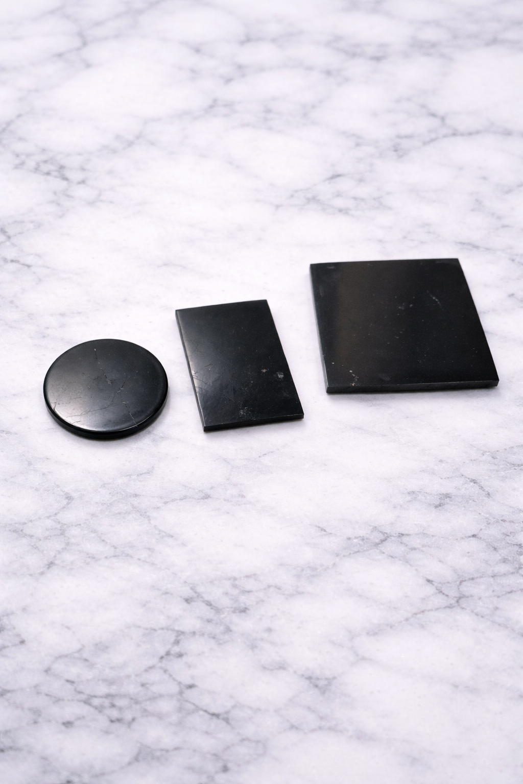 Set of three real shungite phone plates in round, rectangular and square shapes on white marble background