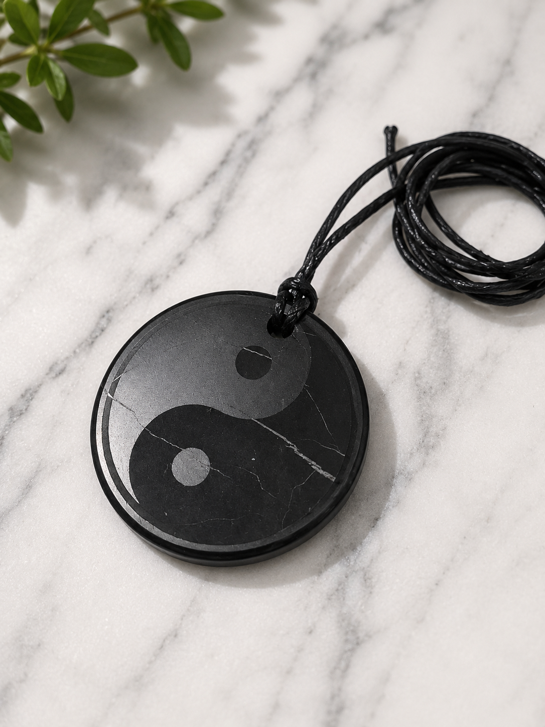 Round shungite yin-yang pendant necklace handcrafted from genuine shungite stone, displayed on a white marble surface with soft natural lighting. The polished black pendant features subtle natural mineral veining and hangs from a black adjustable cord, creating a balanced and timeless design inspired by the yin-yang symbol and the harmony of opposing forces.