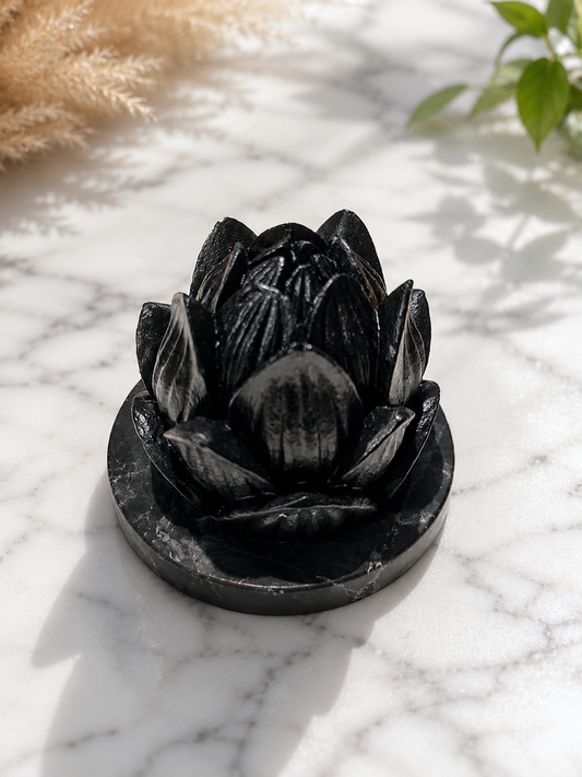 Handcrafted black shungite lotus candle holder on a polished stone base, photographed on a white marble surface with soft natural lighting and botanical accents.