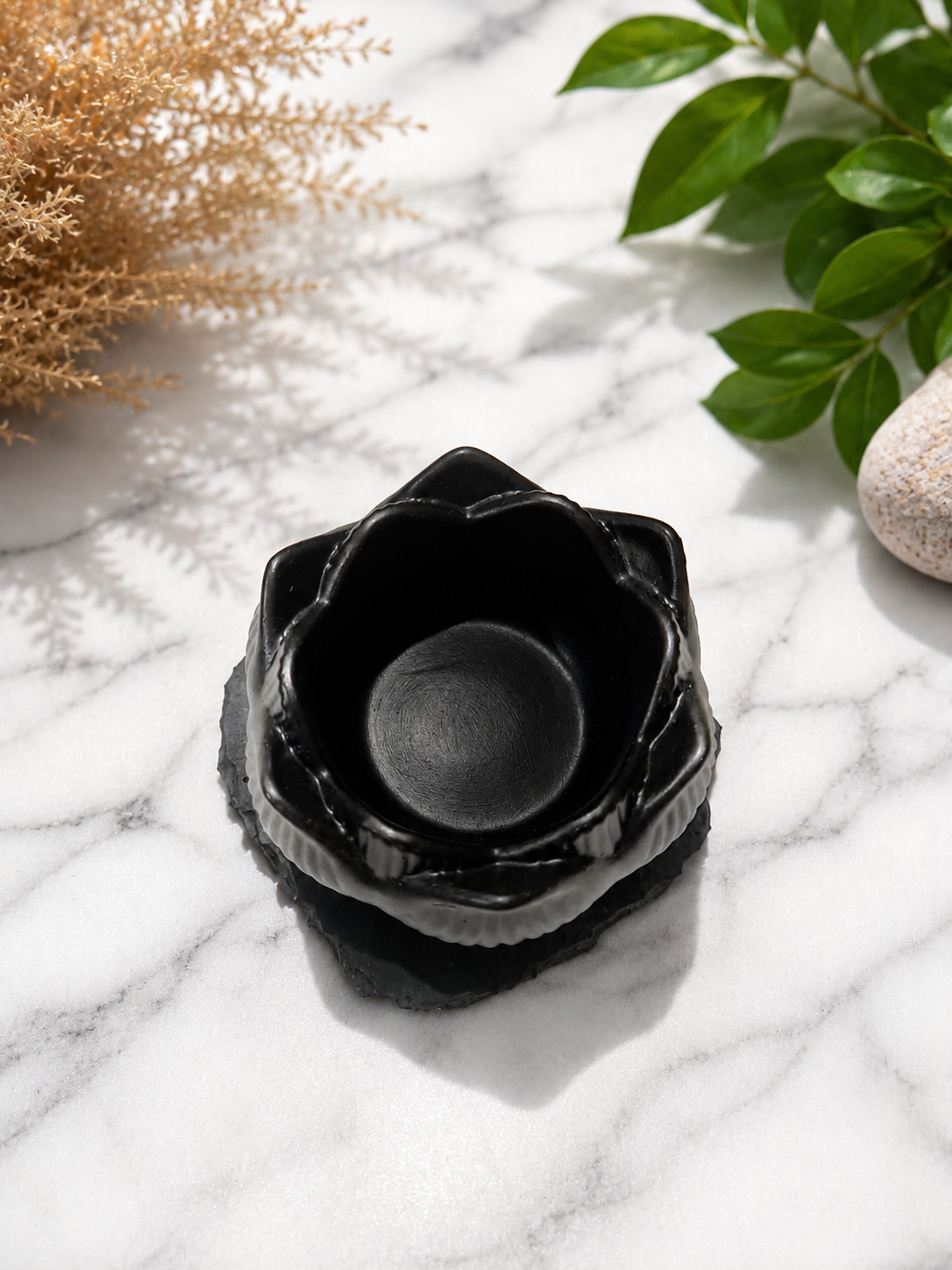 Black shungite lotus candle holder bowl displayed on a marble surface with soft natural lighting, gentle shadows, and calming botanical accents in a minimalist spa-inspired setting.