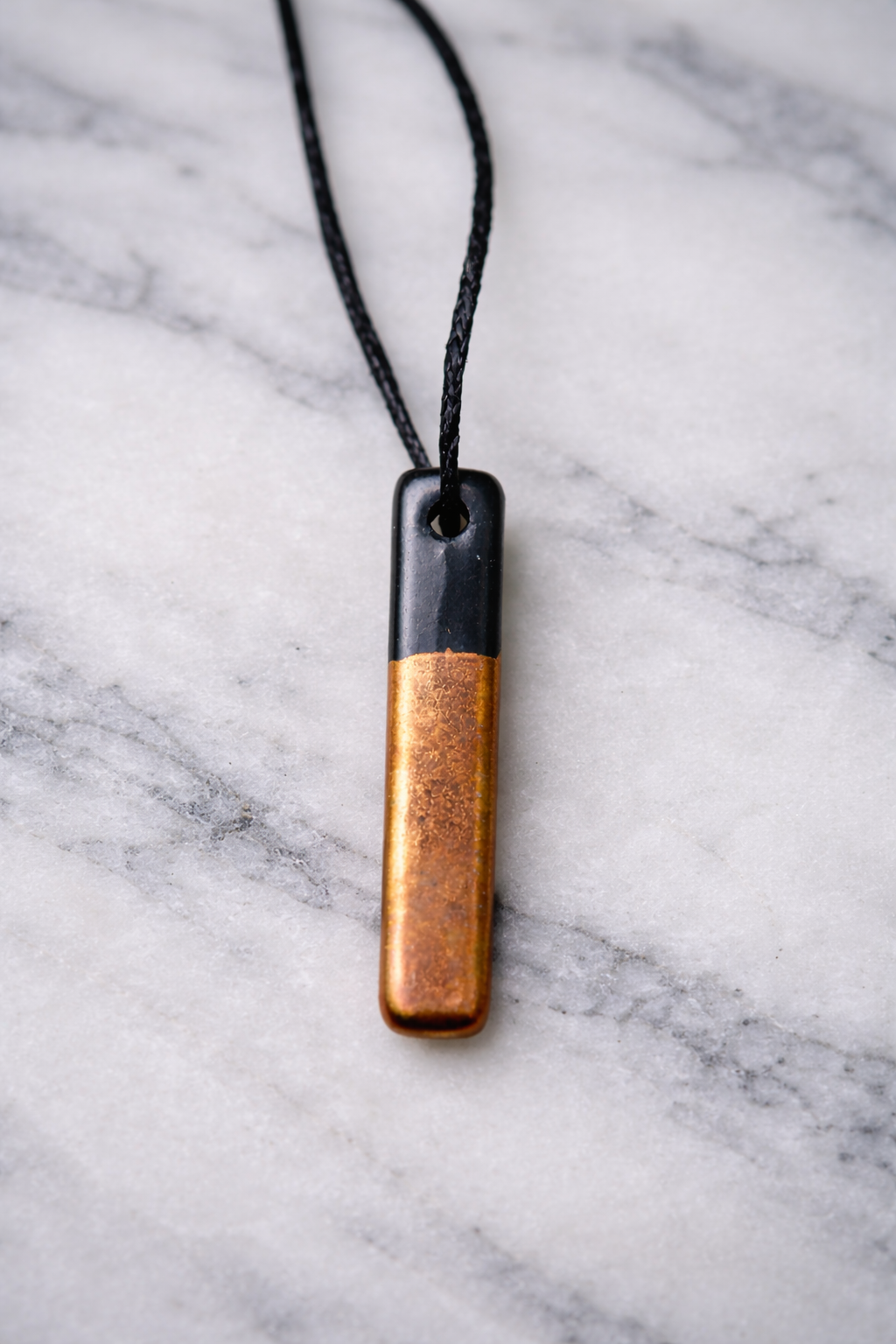 “Two-tone shungite and copper pendant hanging on black cord against smooth marble background”