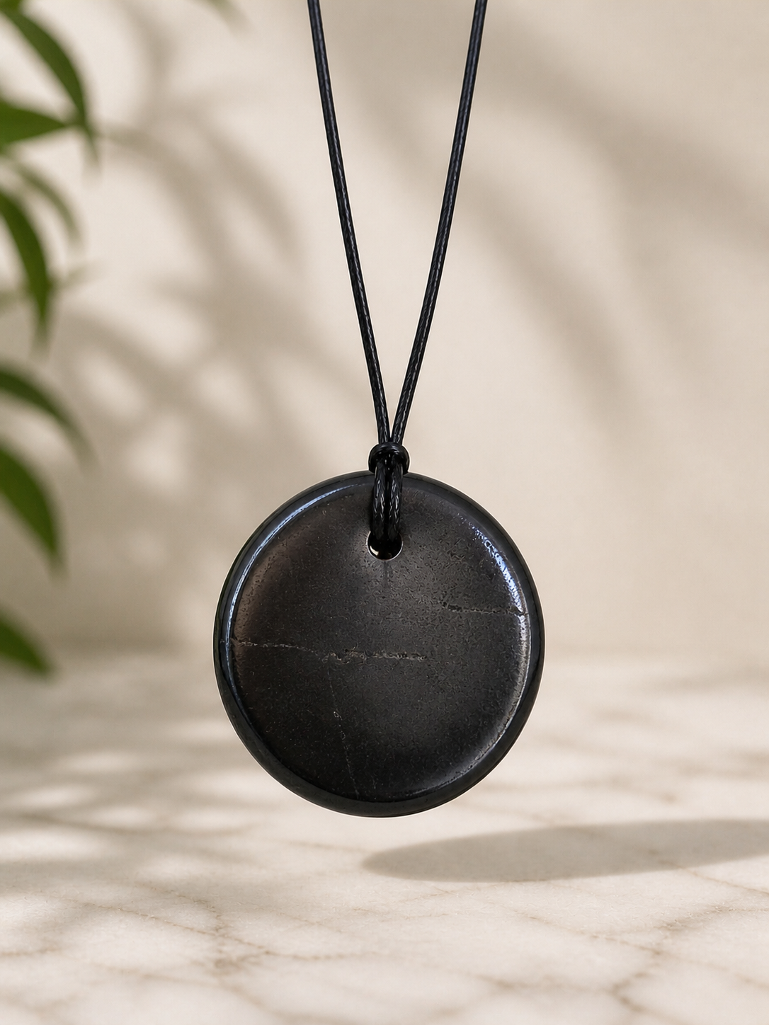Round shungite pendant necklace hanging on a black cord above a marble surface, featuring smooth polished stone texture, soft natural lighting, and minimal earthy background.