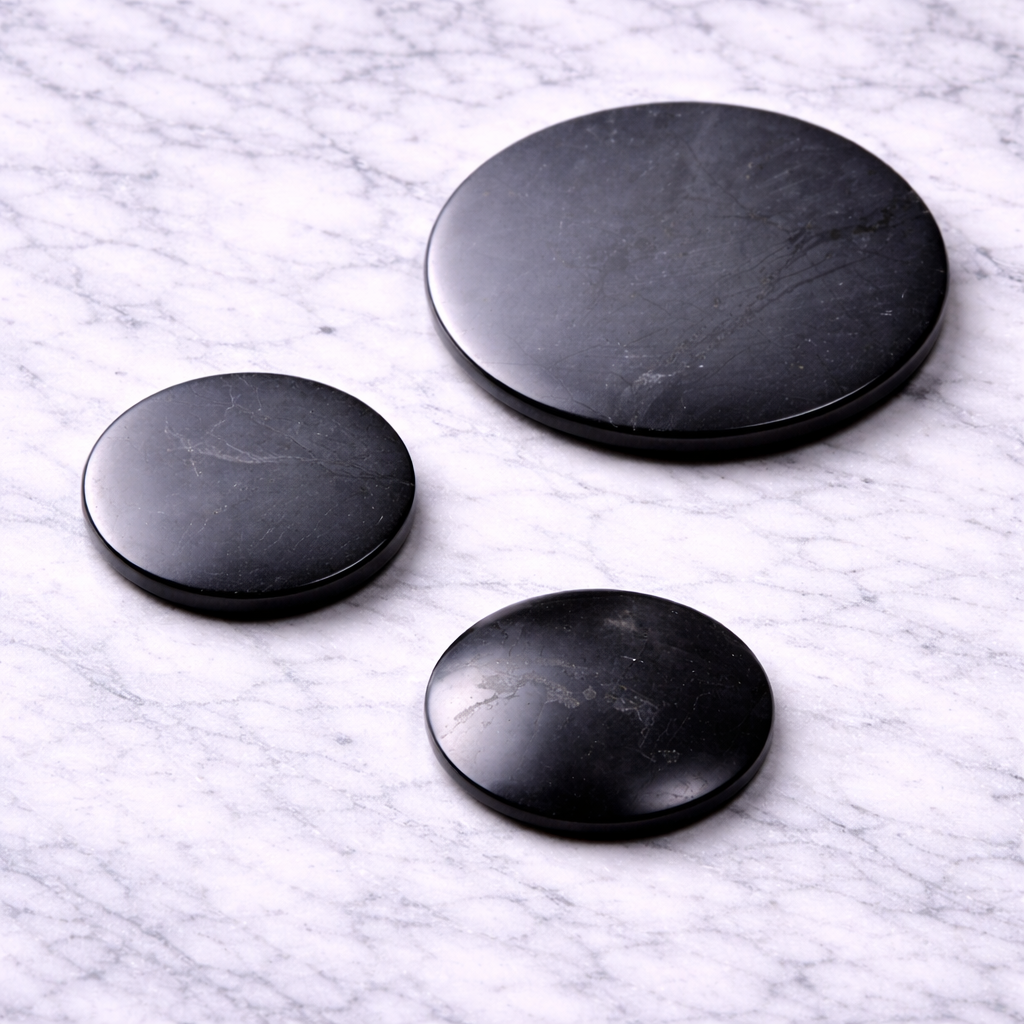 Three black shungite EMF protection phone plates in different sizes (50mm, 30mm flat, and 30mm convex) displayed on a white marble surface, showing smooth polished finish for attaching to electronic devices.