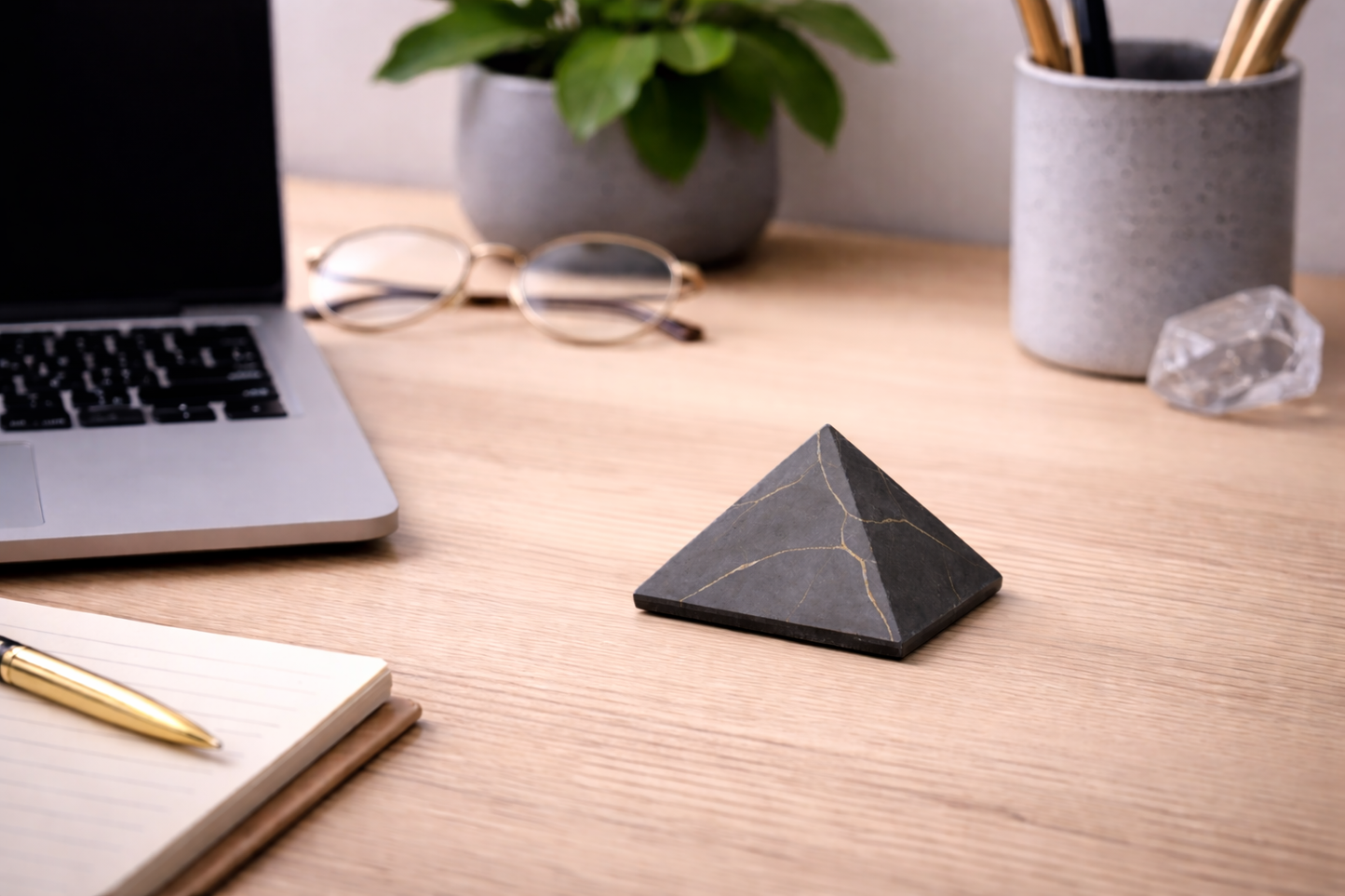 Small matte black shungite pyramid with subtle natural veins sitting on a light wooden desk beside a laptop, notebook, glasses, crystal, and potted plant, styled as a minimalist workspace setup.