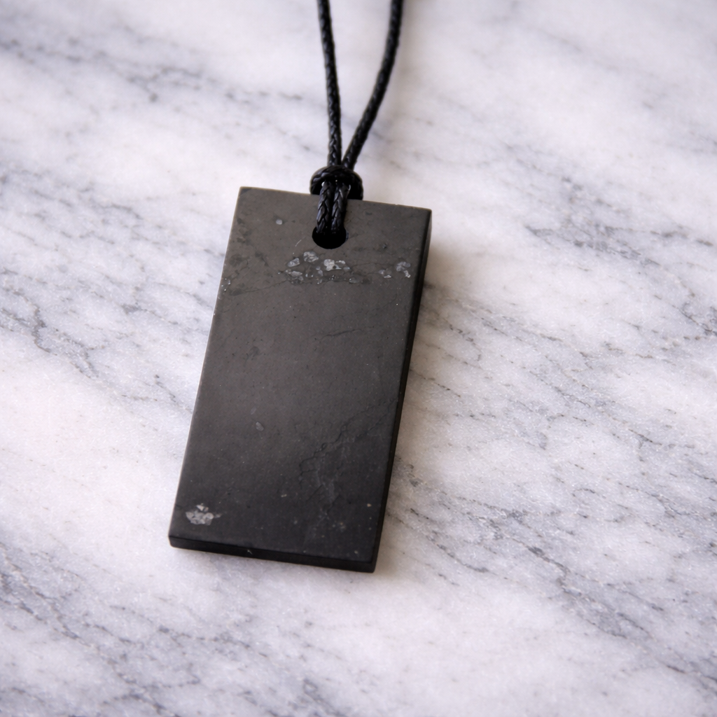 Rectangular shungite pendant on a black cord resting on a white marble surface, showing natural silver mineral flecks and unique stone markings.
