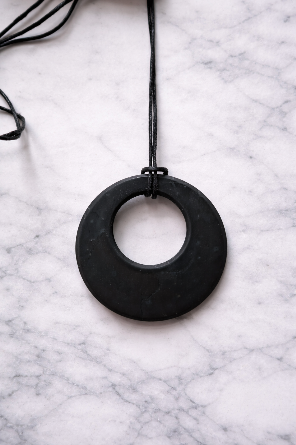 Round shungite pendant necklace on black cord displayed on white marble background