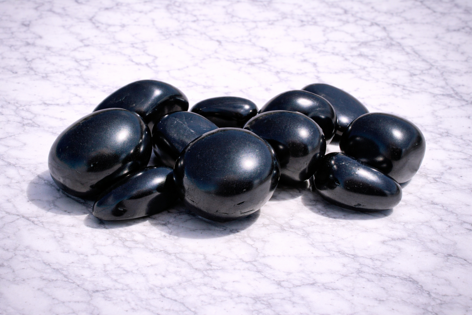 Scattered polished shungite stones on a white marble surface, showing smooth black tumbled crystals with a glossy natural shine.