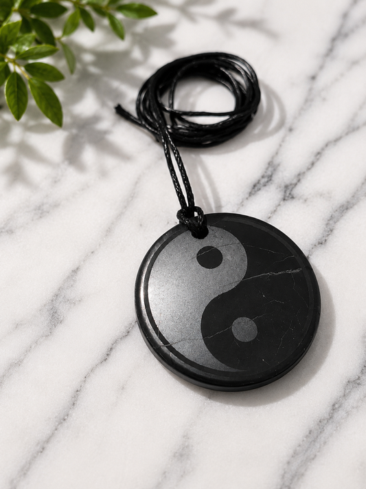 Round yin-yang shungite pendant necklace handcrafted from genuine shungite stone, displayed on a white marble surface with soft natural lighting. The polished black pendant features subtle natural mineral veining and hangs from a black adjustable cord, symbolizing balance, harmony, and the union of opposing forces.