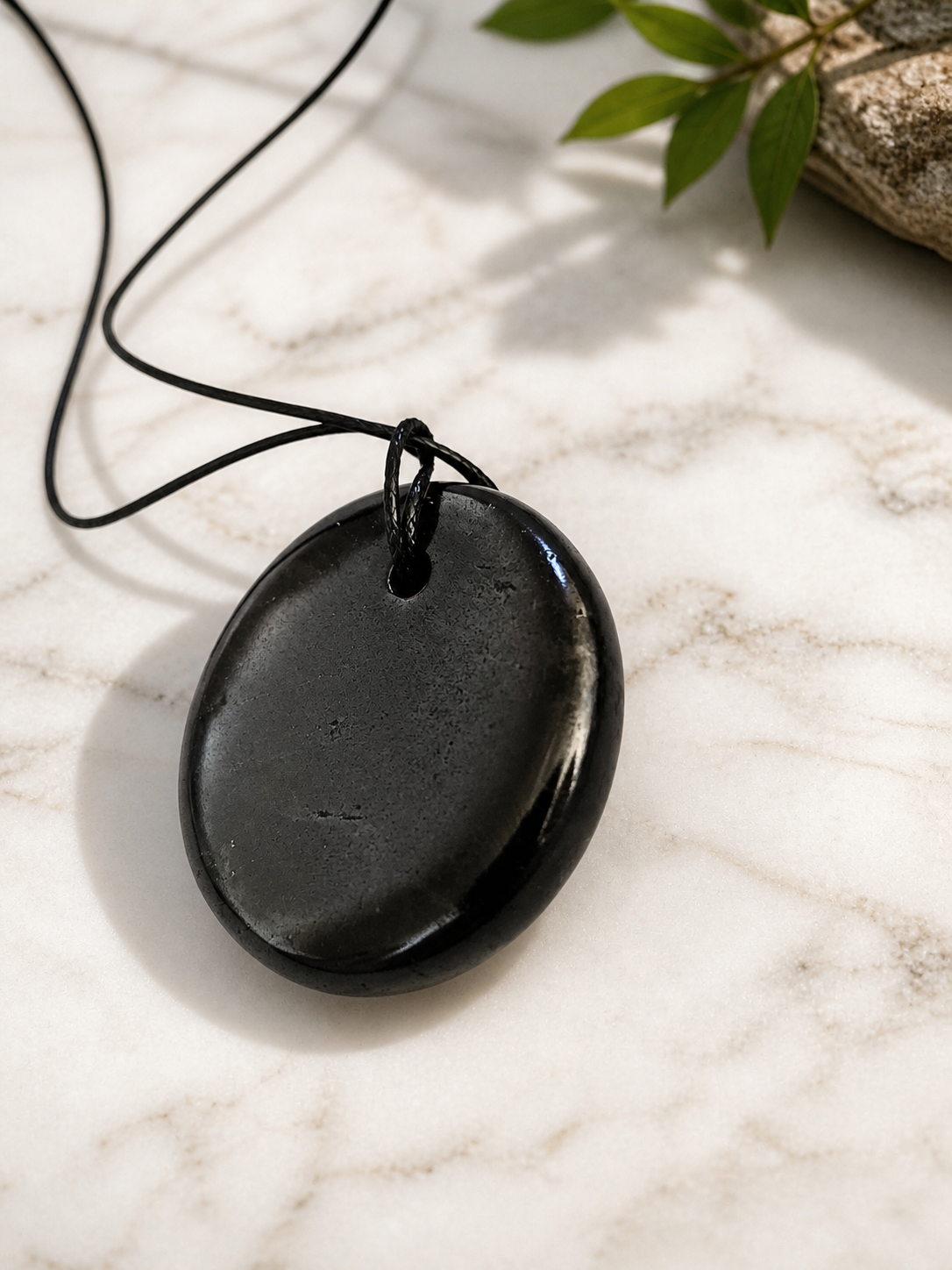 Polished round shungite pendant necklace on a light marble surface, showing smooth natural stone texture with soft reflections, minimal styling, and gentle natural lighting.