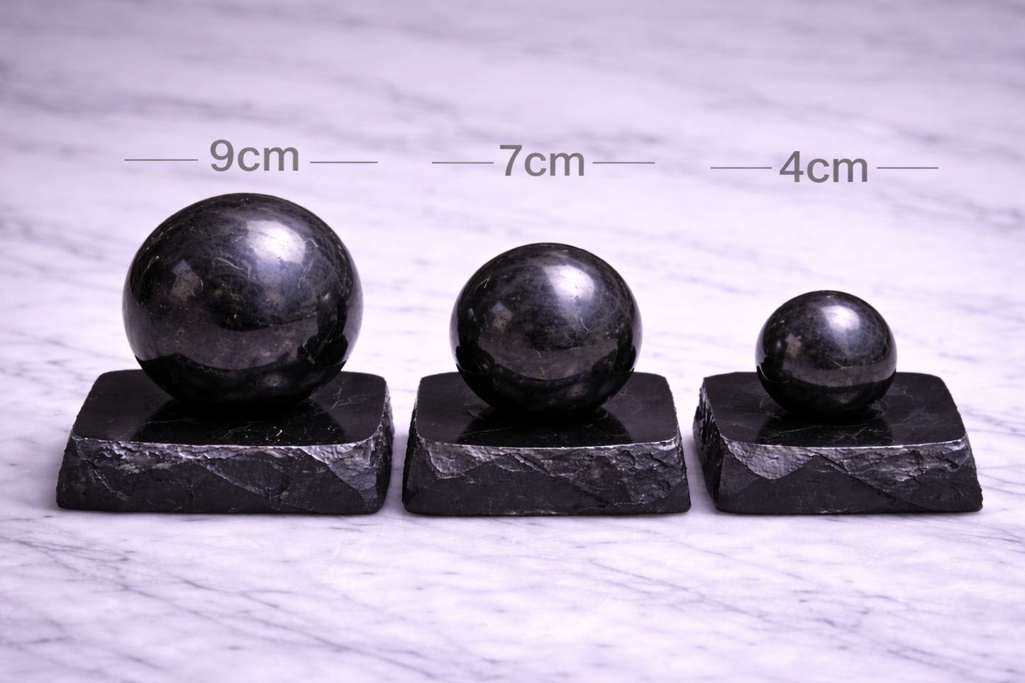“Three polished shungite spheres on individual stands labeled 9cm, 7cm, and 4cm on marble surface”