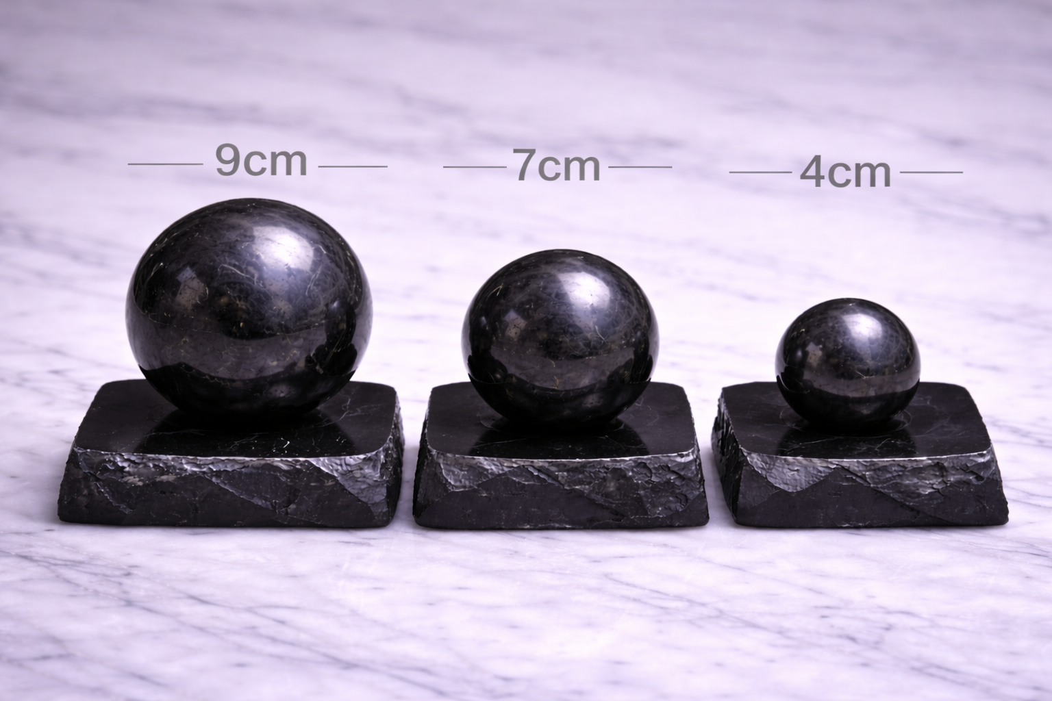 “Three polished shungite spheres on individual stands labeled 9cm, 7cm, and 4cm on marble surface”