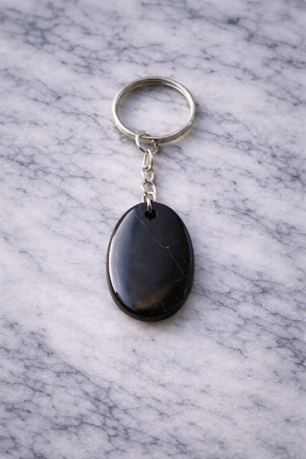Polished oval black shungite keychain with silver chain and ring, displayed on white marble surface with soft gray veining.