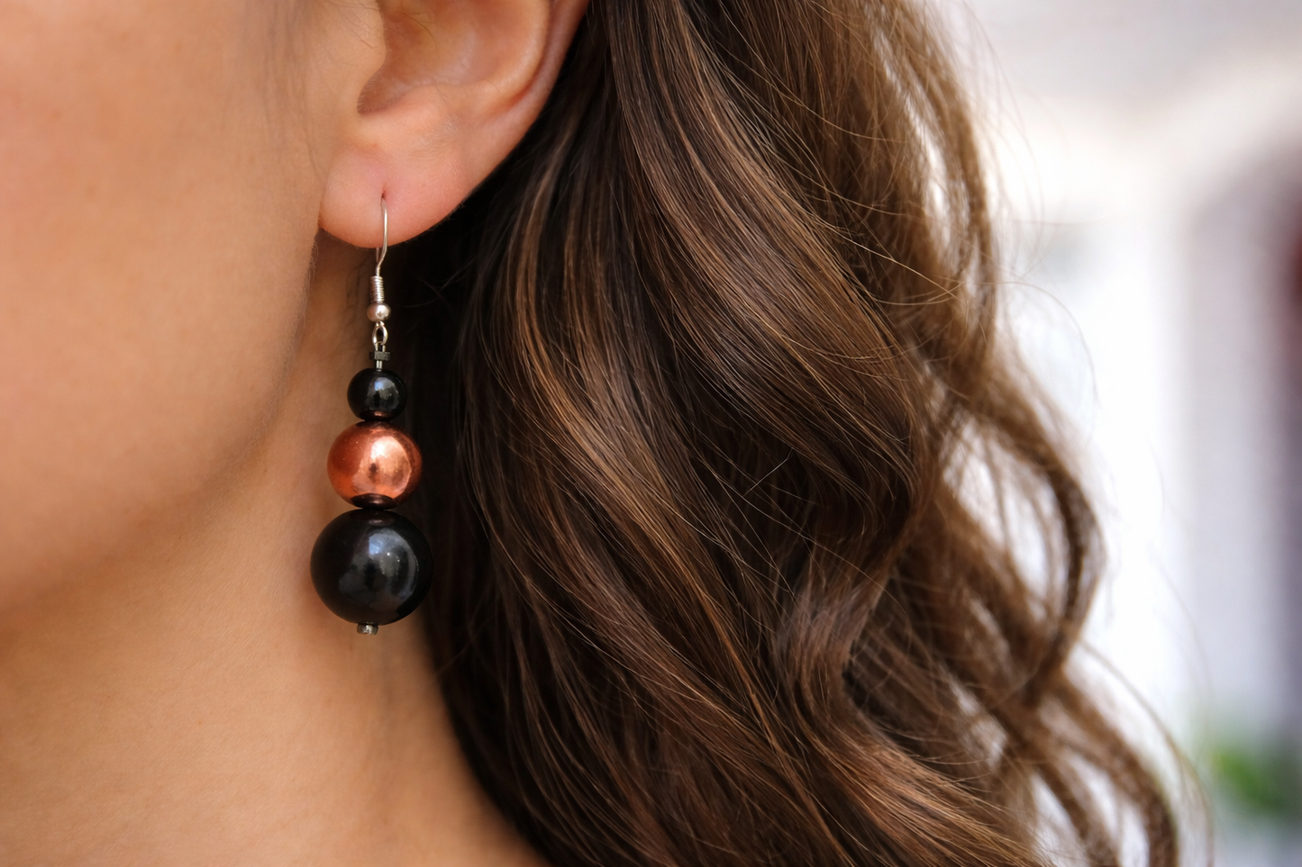 Close-up of a woman’s ear wearing handmade drop earrings with black shungite beads and a copper bead on silver hooks, framed by long wavy brown hair and softly lit natural lighting.